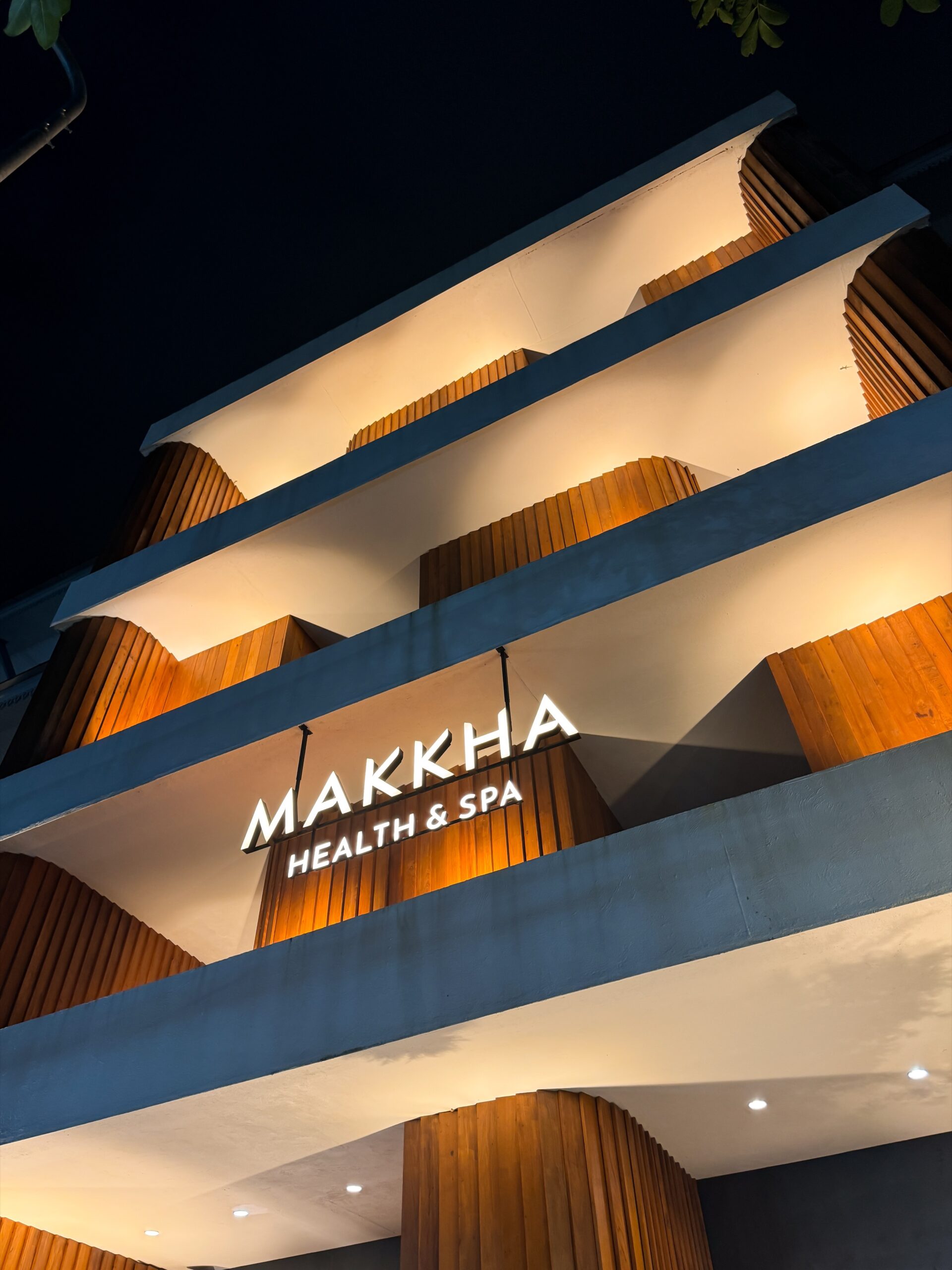 MAKKHA Health & Spa sign illuminated at night on a modern building facade featuring curved concrete balconies and warm wood paneling.