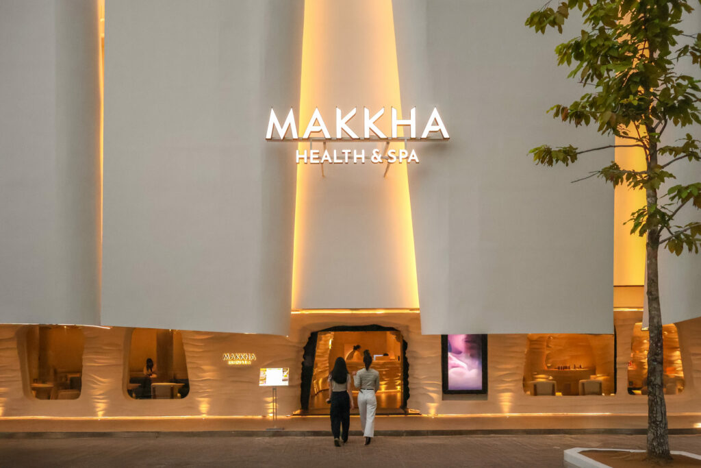 Illuminated MAKKHA Health & Spa exterior featuring a modern white facade, warm golden accent lighting, and two women entering the facility.