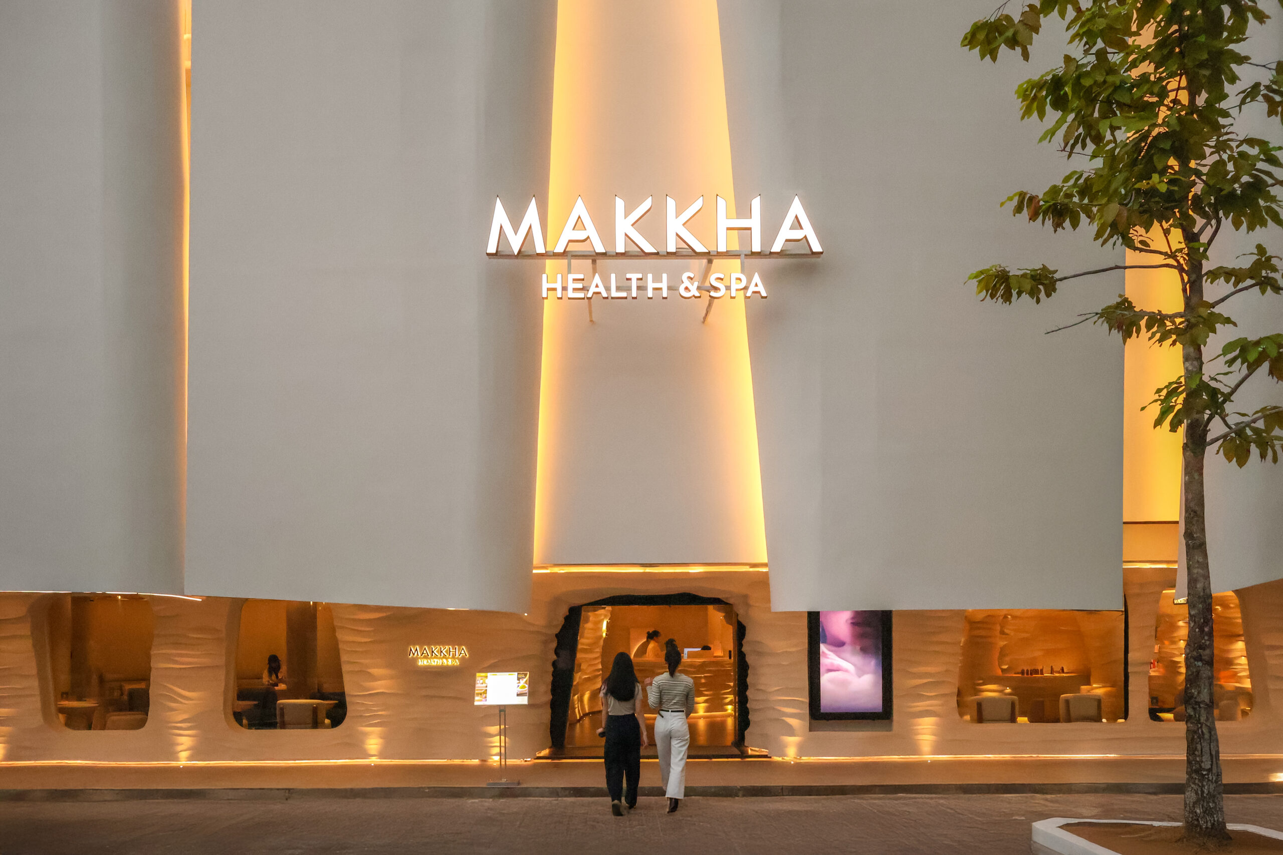 Illuminated MAKKHA Health & Spa exterior featuring a modern white facade, warm golden accent lighting, and two women entering the facility.